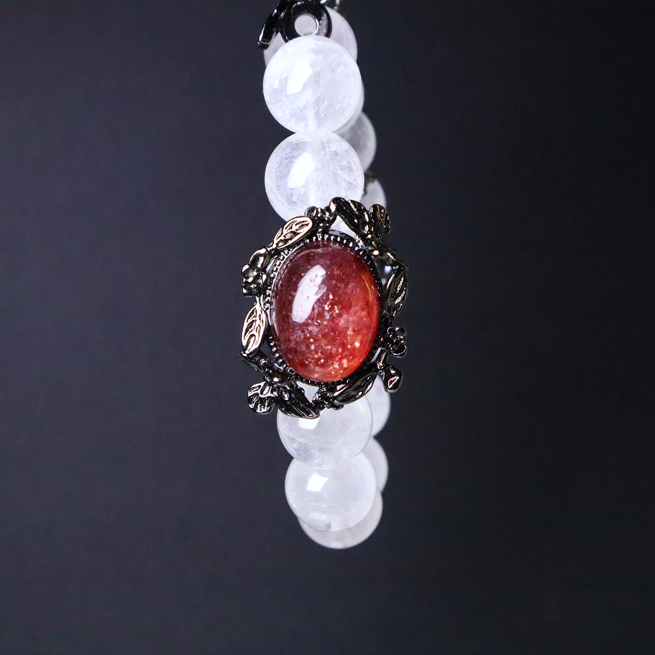 Close-up of sunstone centerpiece set in an ornate black floral bezel, paired with round white phantom quartz beads on a black background.