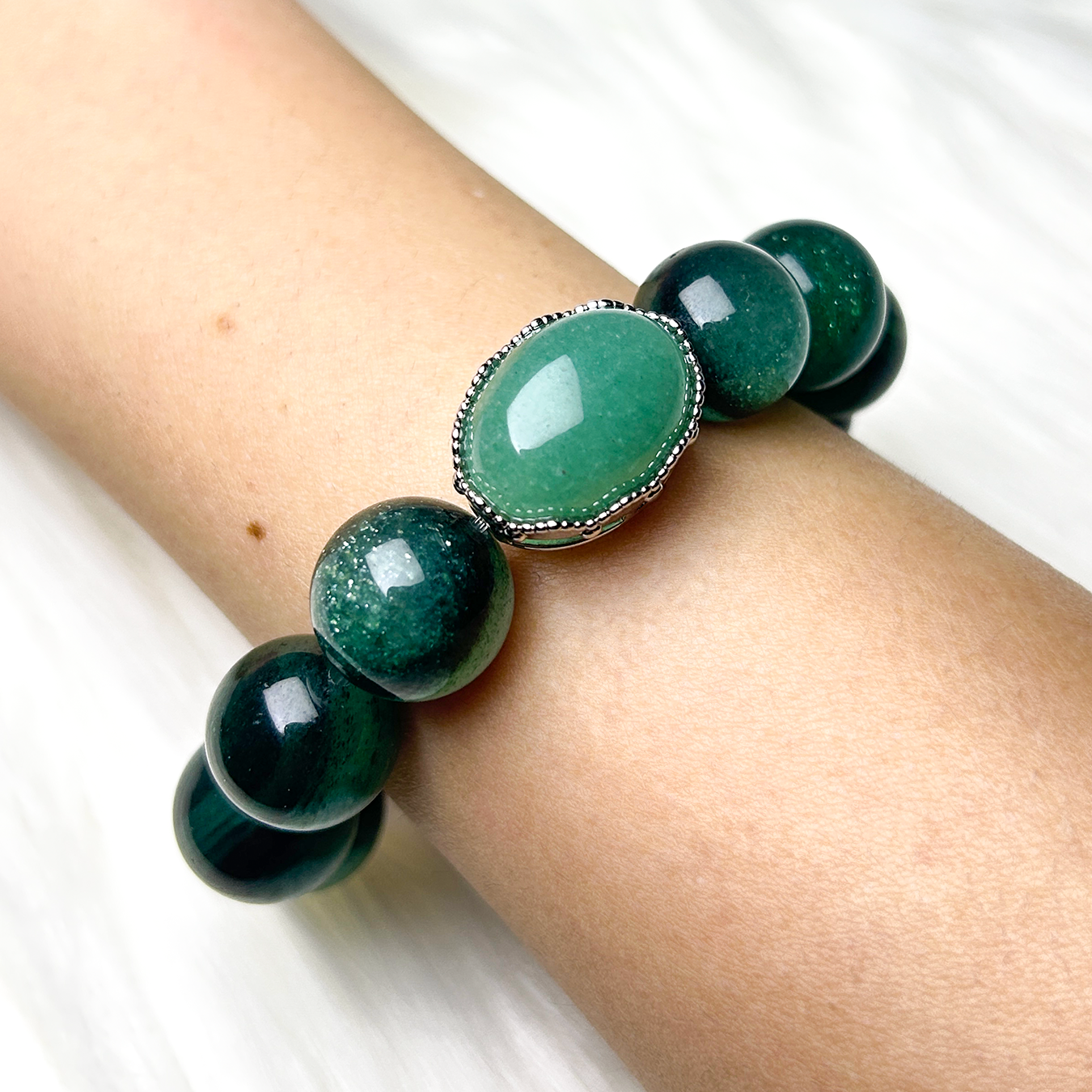 Wrist shot of crystal bracelet featuring a central oval green aventurine centerpiece and glossy green strawberry quartz beads, radiating a calming shimmer on a white fur background.