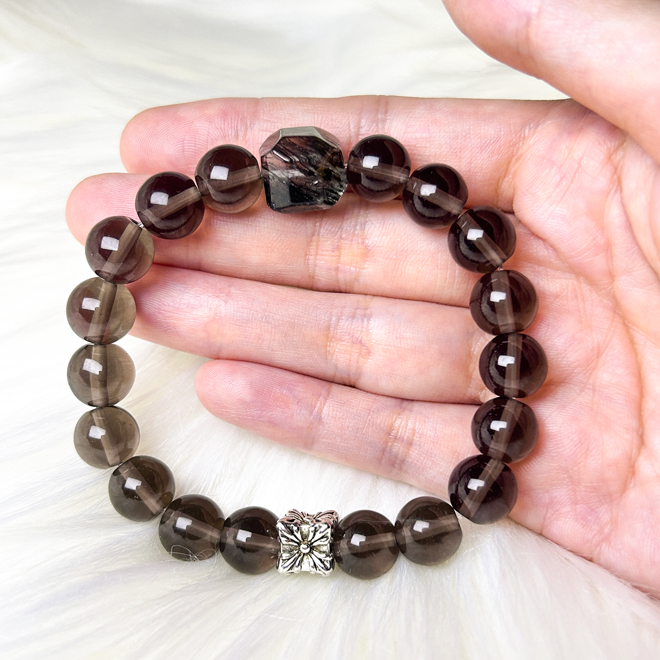 Smoky quartz and rutilated quartz beaded bracelet displayed on hand, showing the gemstone’s natural shine and silver flower charm design.