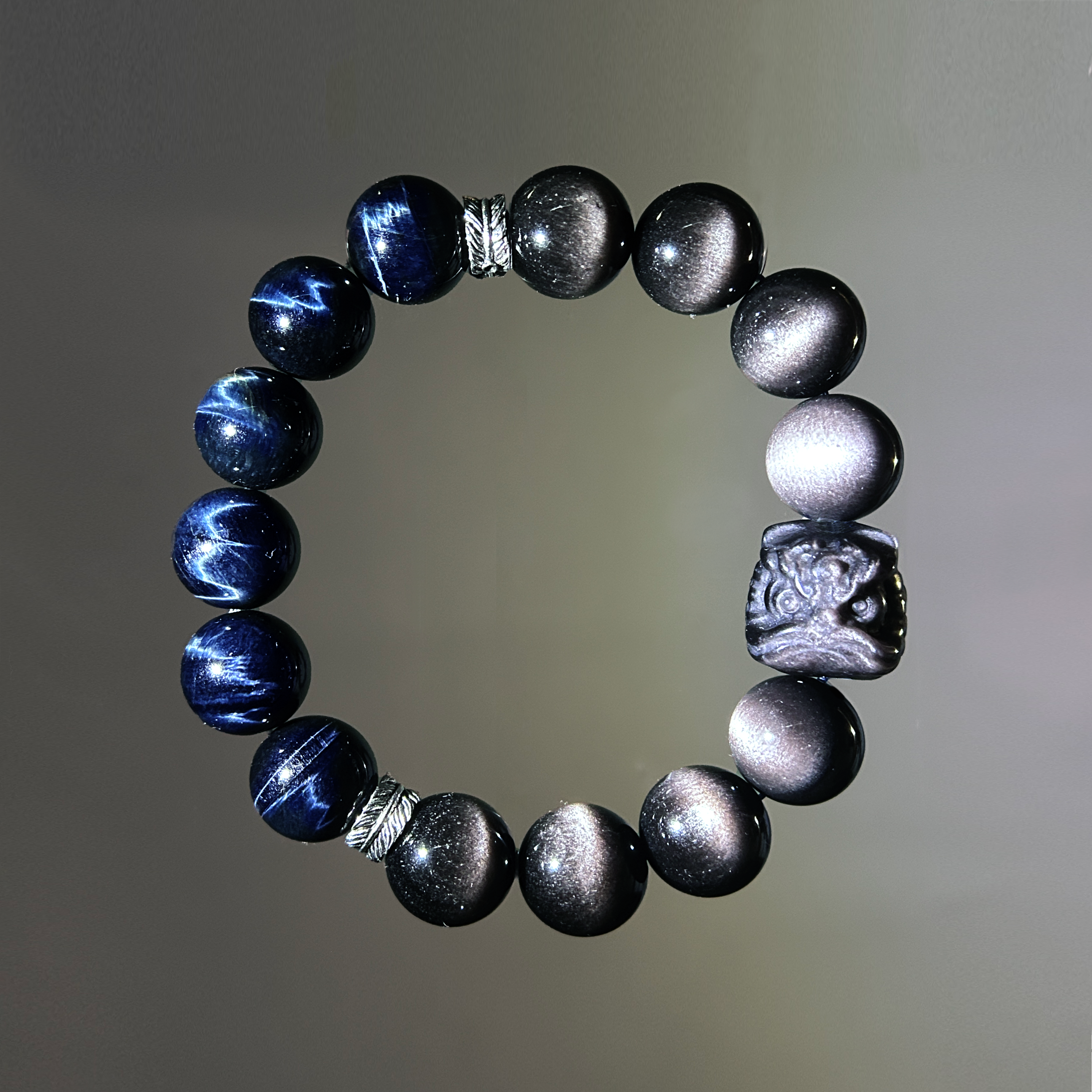 hang lay out of a men bracelet handmade with blue tiger's eye and black obsidian, featuring a pi xiu centerpiece and silver feather spacers in a dark background