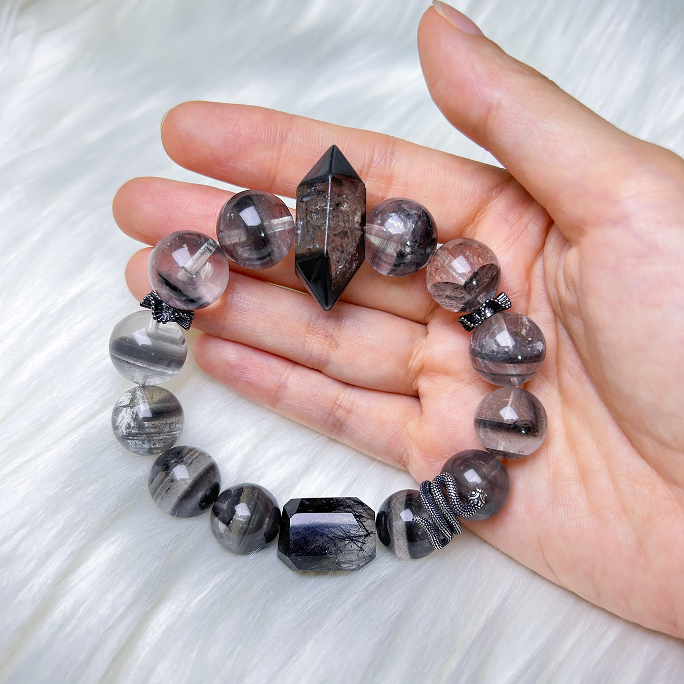 Hand-holding shot of the bracelet with red book background, showcasing bold black phantom quartz tones, faceted centerpiece, and coiled snake charm.