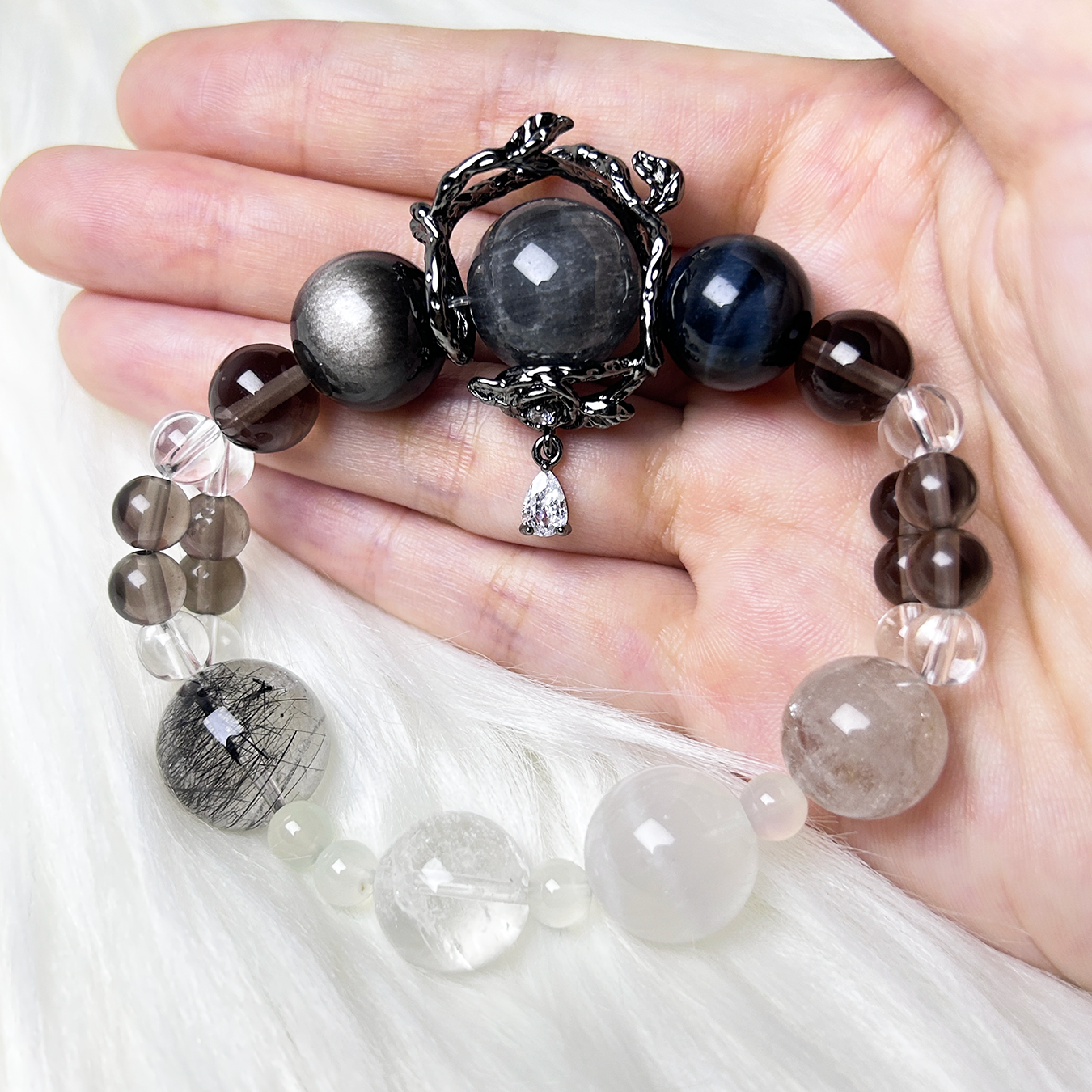 A mixed crystal bracelet with a black vine charm and teardrop crystal pendant is showcased across a hand with elegant neutral-toned beads.
