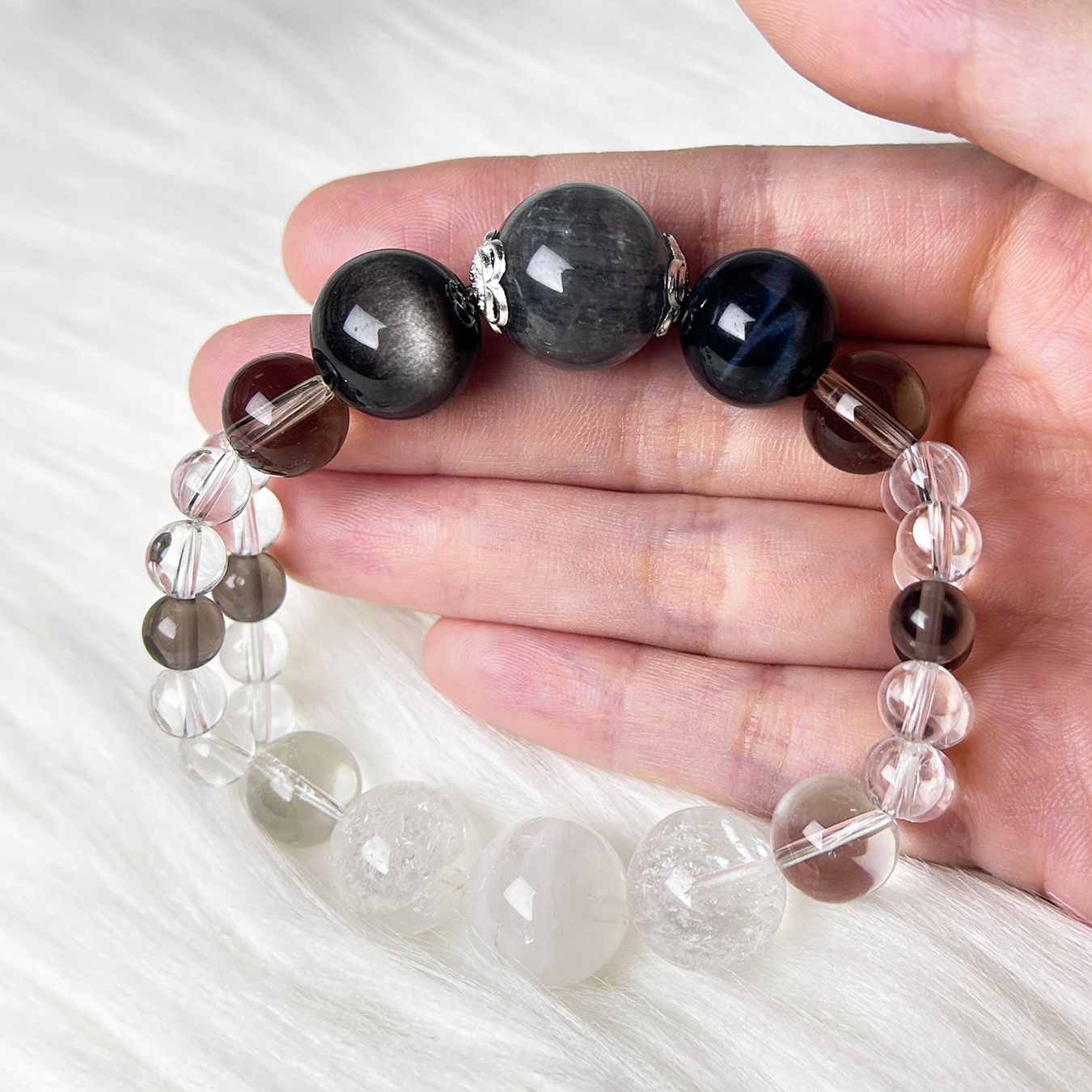 A mixed crystal bracelet featuring earth tone crystal beads, and clear Quartz is gently held in a hand over a white fur background.
