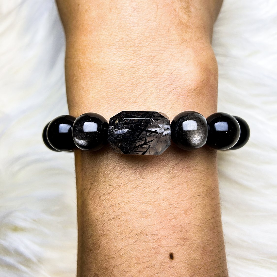 Close-up wrist shot of a handcrafted bracelet featuring a faceted black rutilated quartz centerpiece, surrounded by glossy black obsidian beads, set against a white fur background.