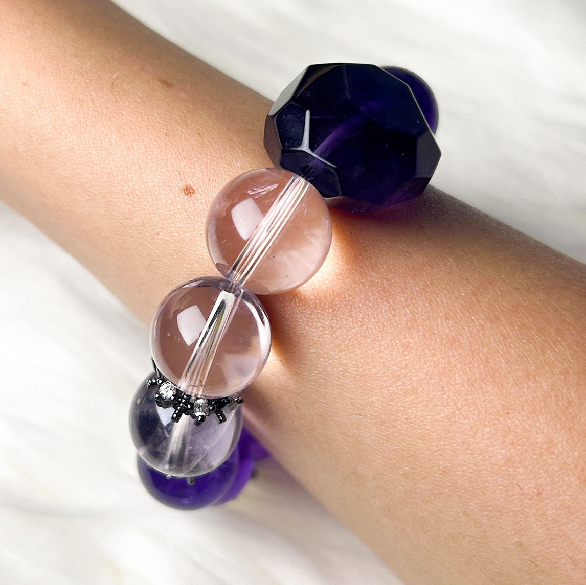 Wrist shot of amethyst bracelet showcasing light pinkish amethyst beads transitioning into a dark faceted amethyst centerpiece, accented with black snowflake spacers.